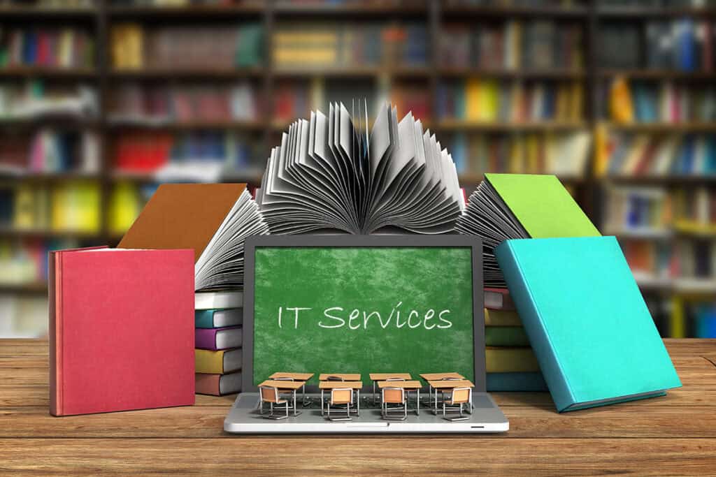 A laptop is on a desk surrounded by books. The screen is showing a green chalkboard with the text "IT Services" written in chalk.