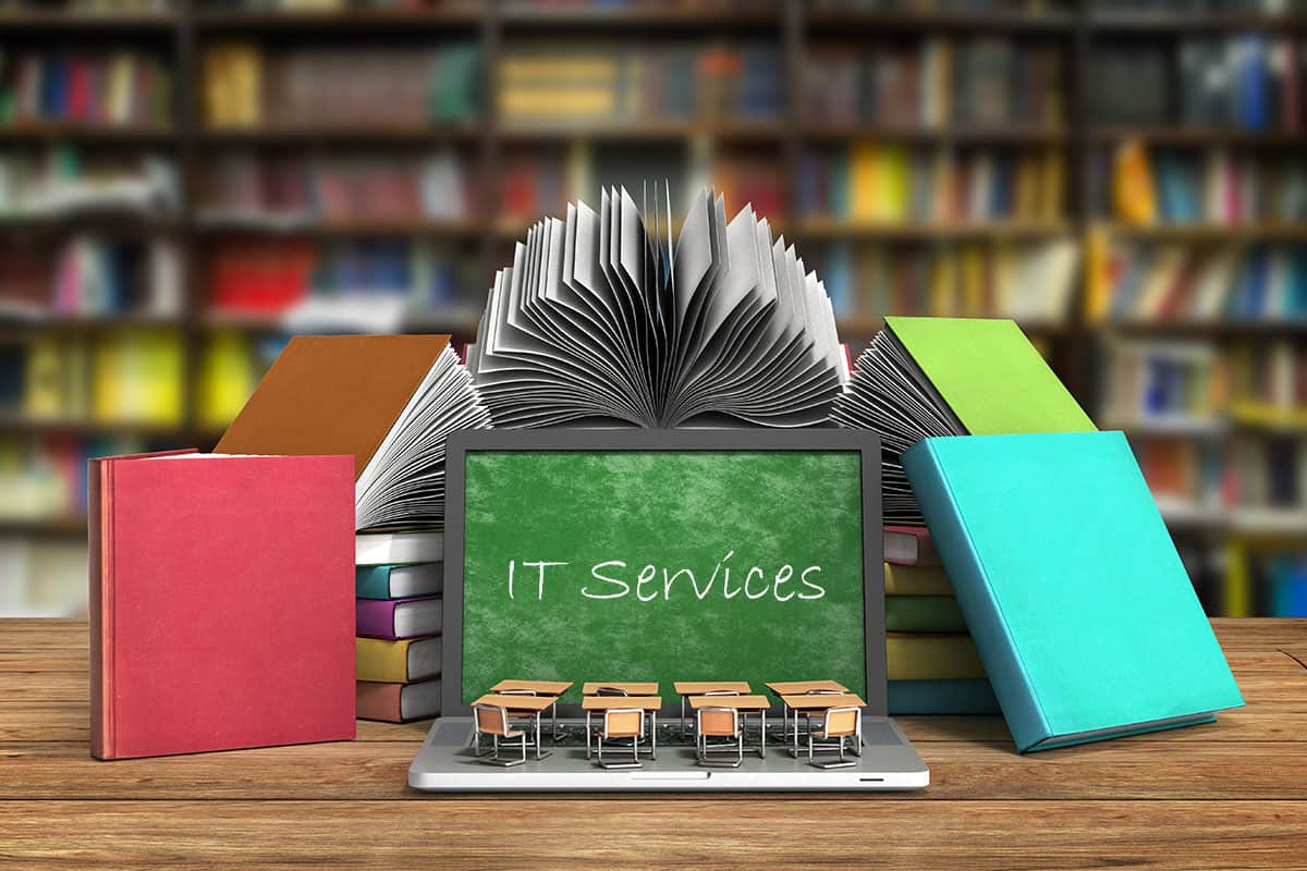 A laptop is on a desk surrounded by books. The screen is showing a green chalkboard with the text "IT Services" written in chalk.