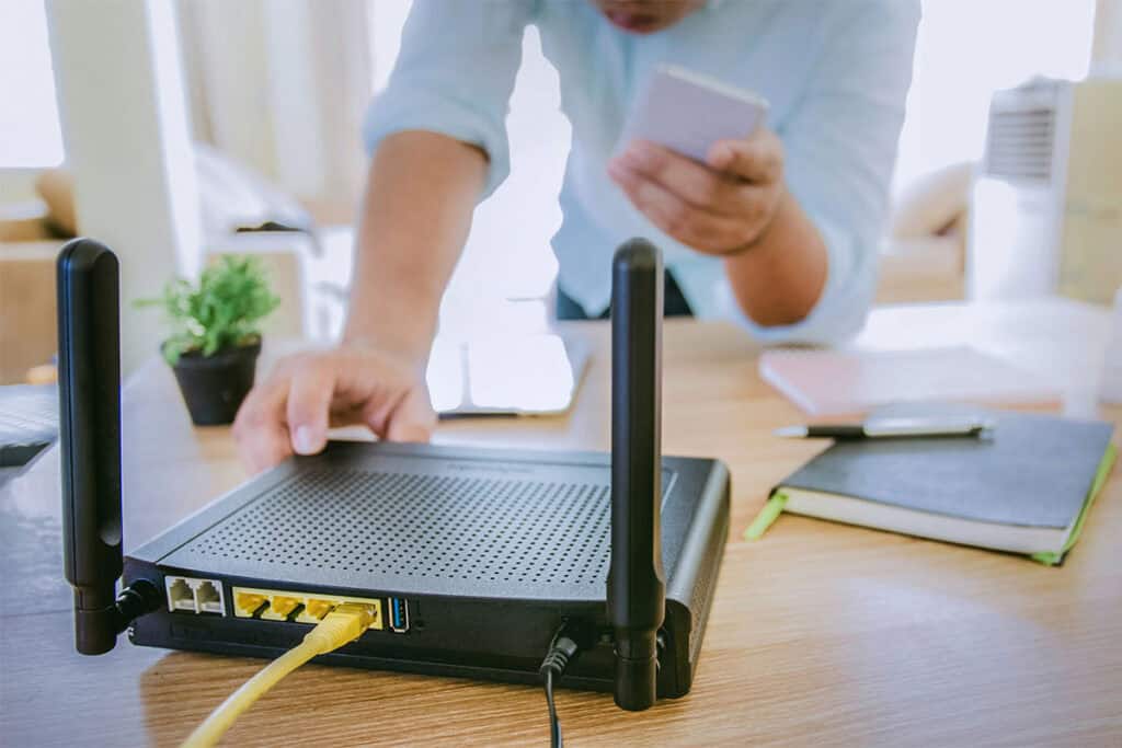 A person adjusts a wireless router on a wooden table, connected with an Ethernet cable. They hold a smartphone in their other hand, with notebooks and a plant visible in the background.