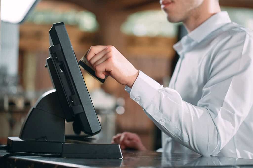 A server wearing a white dress shirt is swiping a credit card through a POS system in a restaurant environment.
